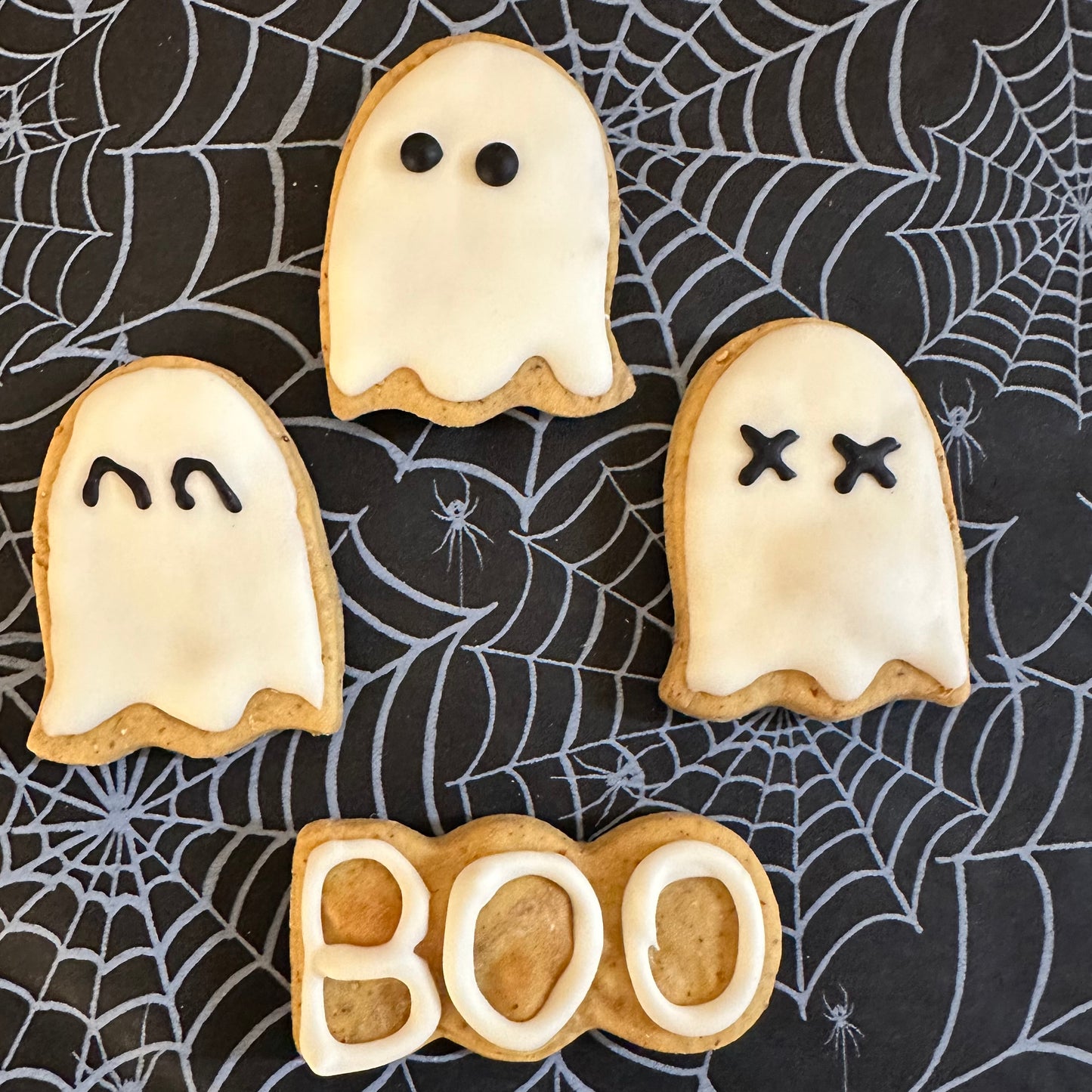 Three ghost-shaped dog biscuits with icing, arranged on a surface with a spider web pattern. One of the ghosts has a smiling face, another has a frowning face, and the third has a 'BOO' text in the same shape.