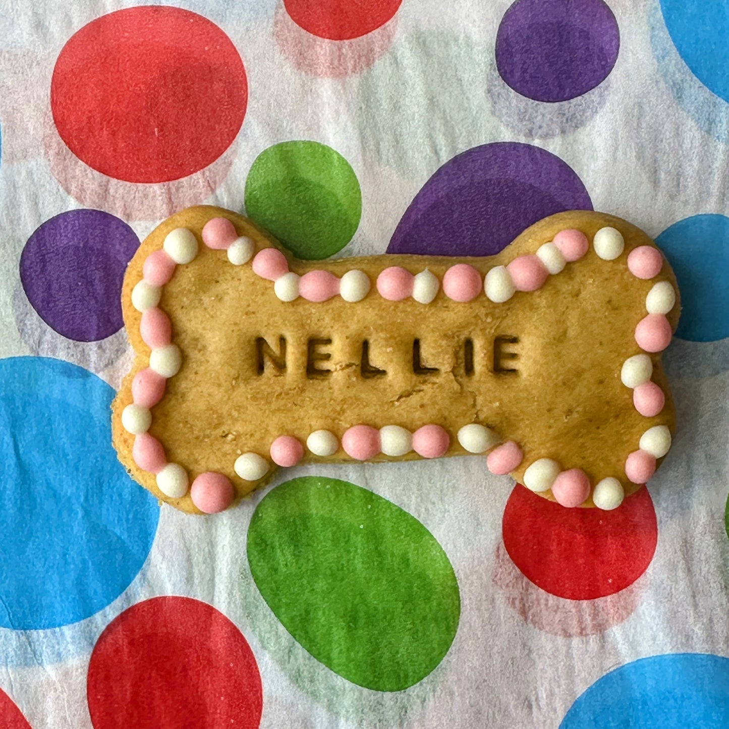 A bone shaped dog biscuit with pink and white dots around the outside with dog name stamped in the middle