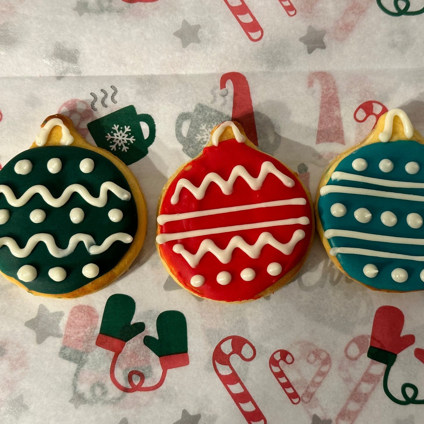 Three decorated dog treats shaped like Christmas baubles, featuring red, green, and blue colors with white and black accents, placed on a background with Christmas-themed pattern.