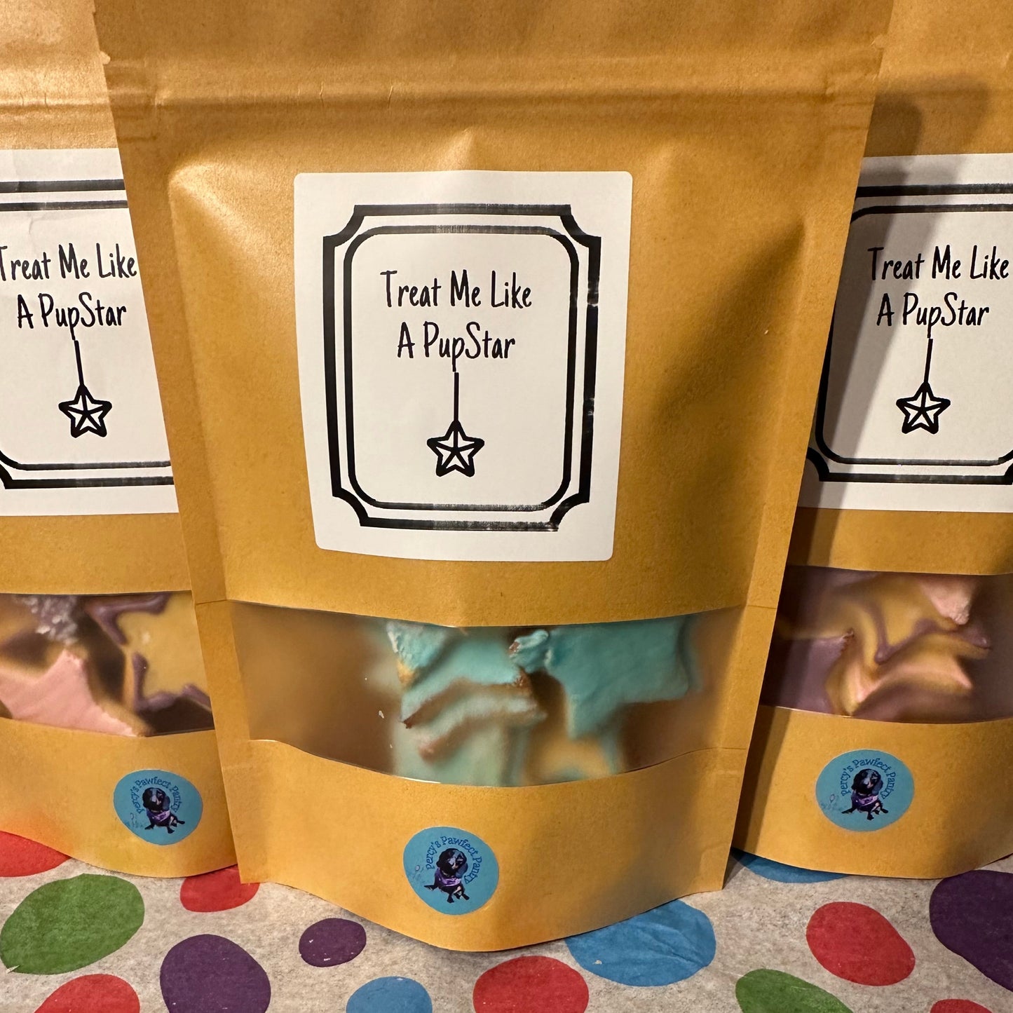 Three golden treat bags with clear windows showing colourful, star-shaped dog treats inside. Each bag has a label with the text 'Treat Me Like A PupStar' and a star symbol.