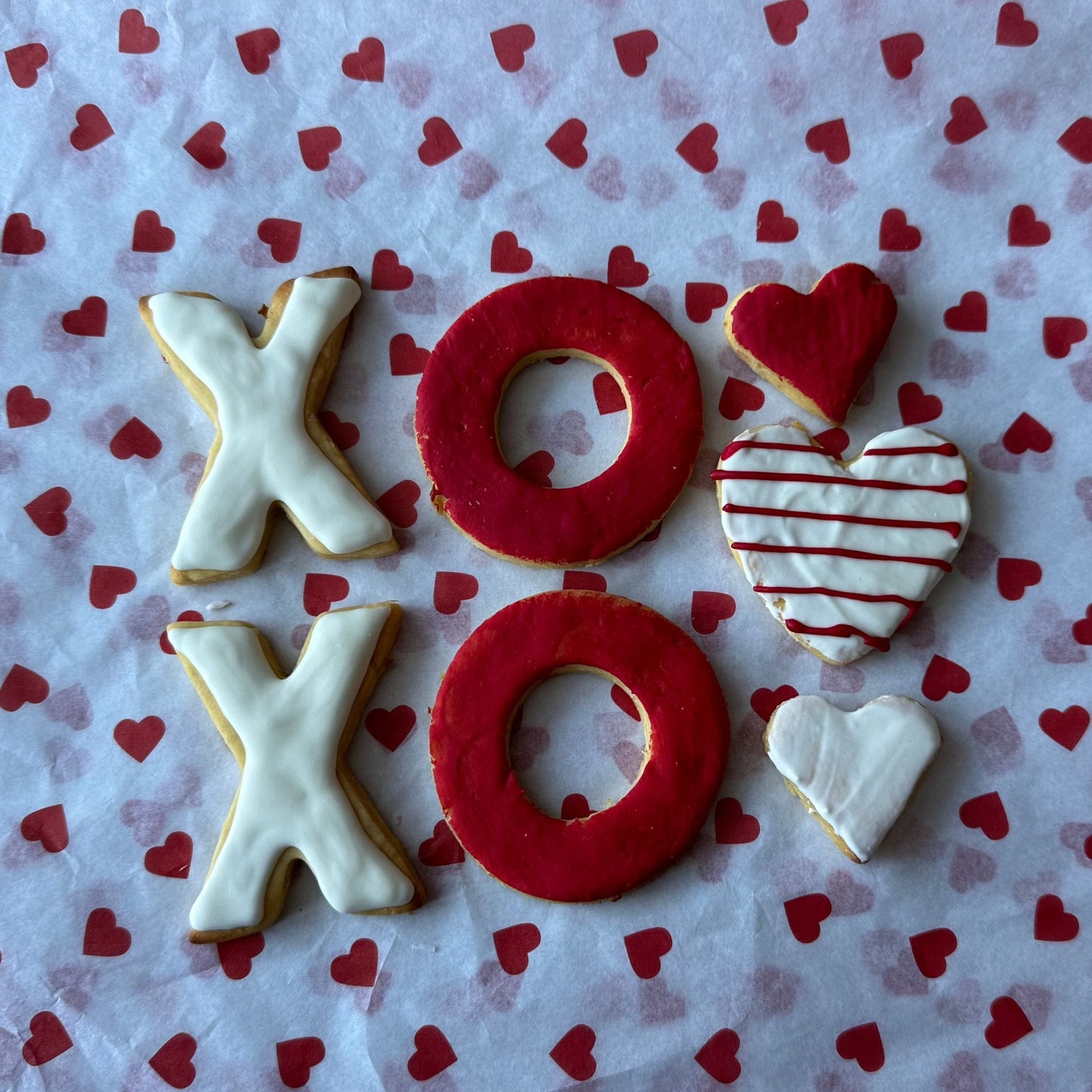 A selection of cookies decorated with icing, spelling out 'XOXO' accompanied by a heart-shaped cookie and two mini heart-shaped treats, presented on a surface with a heart pattern.