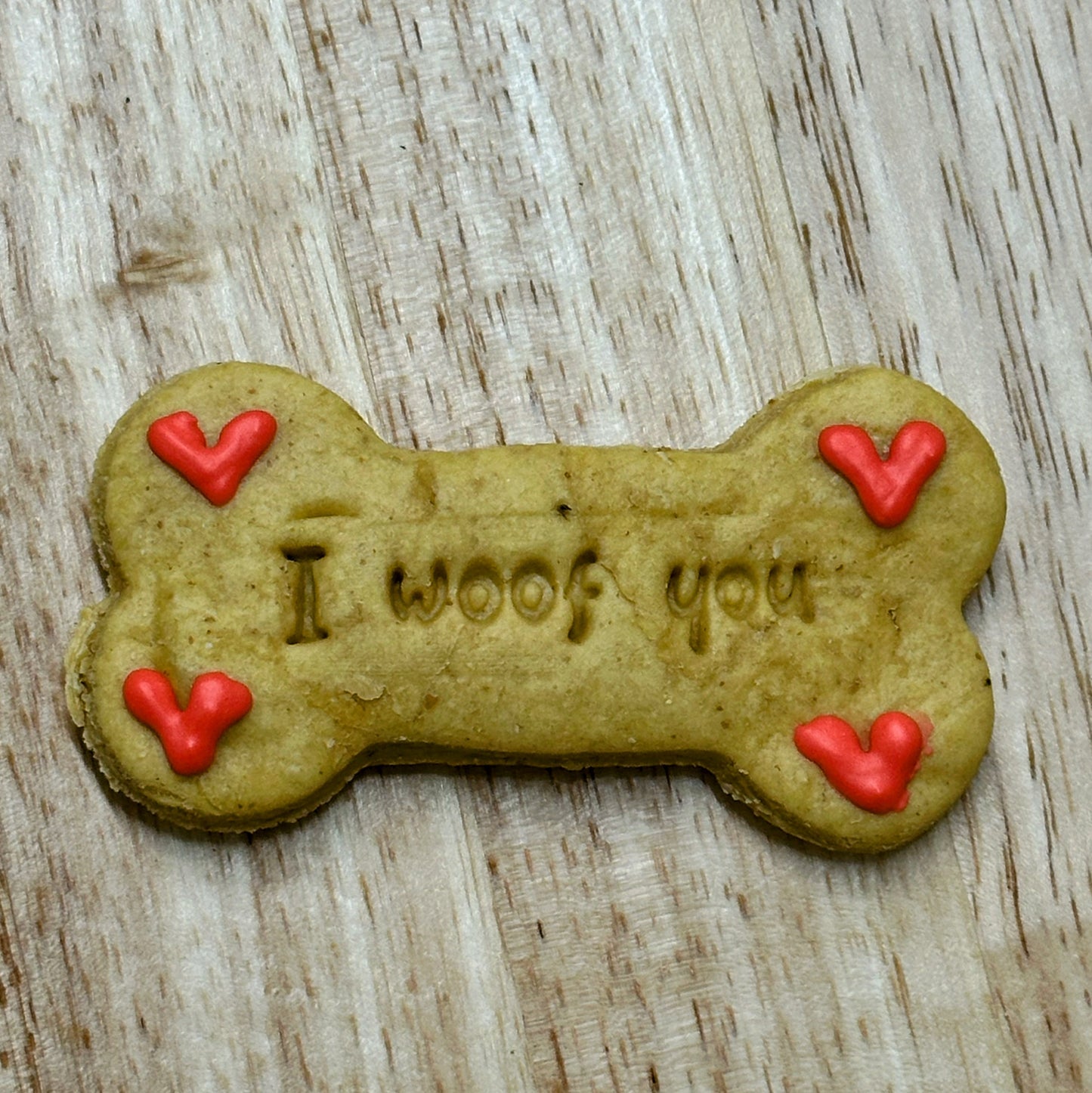 A bone-shaped dog biscuit with 'I woof you' written on it and decorated with red love hearts.