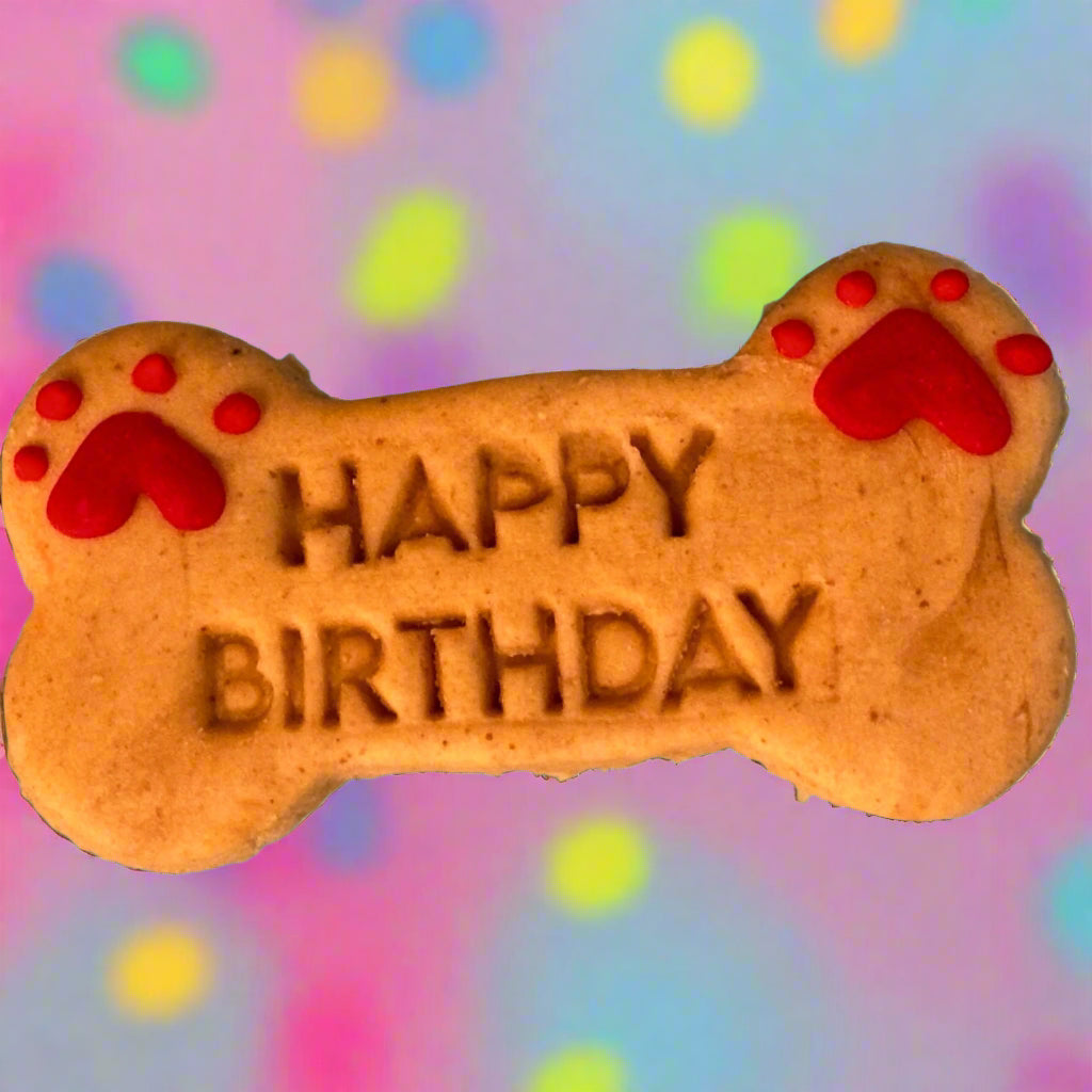 A bone-shaped dog biscuit with 'HAPPY BIRTHDAY' text and red paw prints on it, against a colourful background.