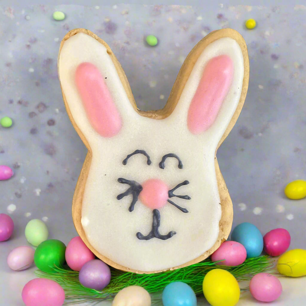A bunny-shaped dog biscuit decorated with dog-friendly icing, presented on a background with colorful Easter eggs.