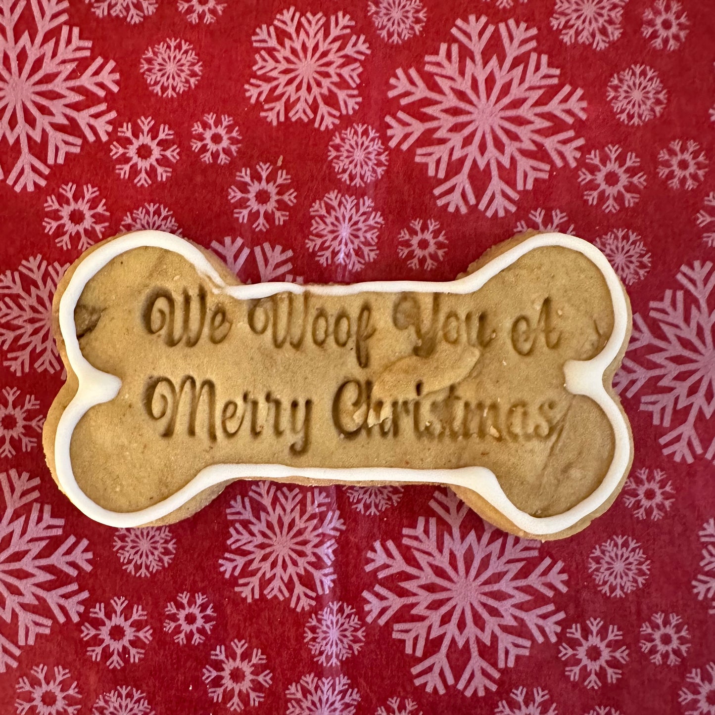 A dog bone-shaped Christmas cookie with the inscription 'We Woof You a Merry Christmas' on a red background with white snowflake pattern.