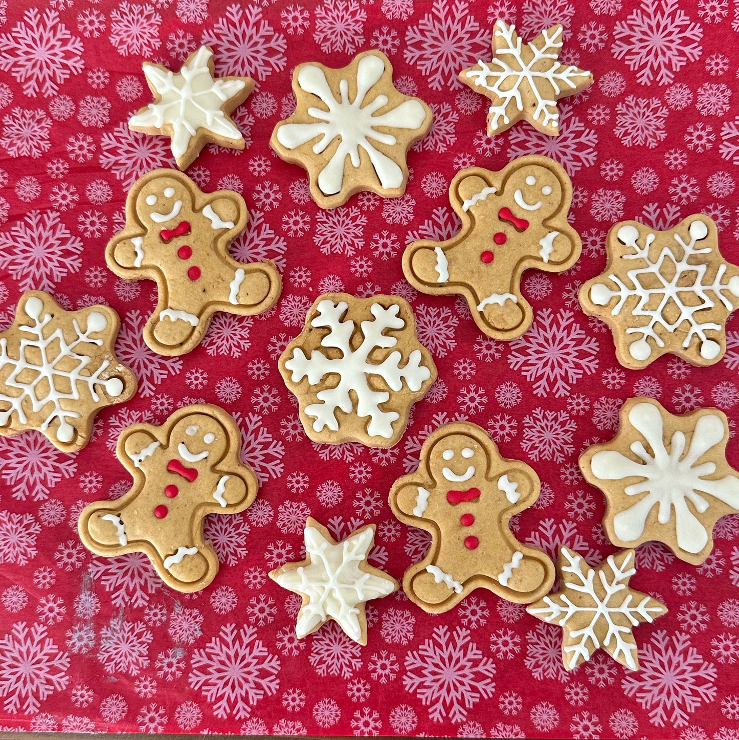 A variety of iced gingerbread man and snowflake-shaped dog treats on a red patterned surface.