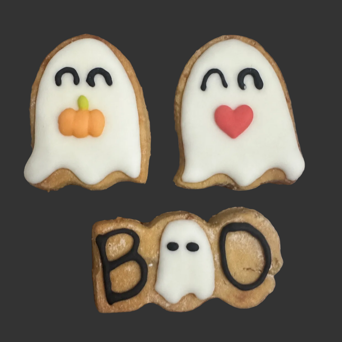 Three decorated Halloween cookies on a wooden surface: two ghost-shaped cookies with white icing, one with a small orange pumpkin and one with a red heart, plus one long cookie spelling BOO with a small ghost in the centre.