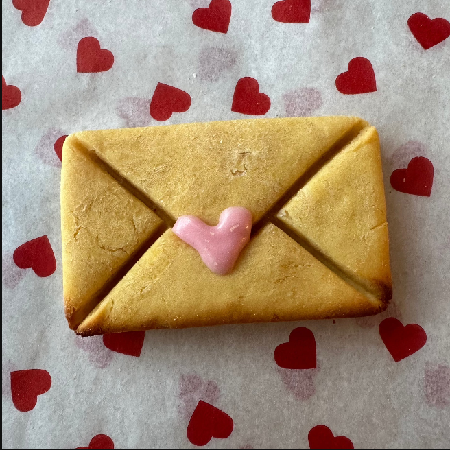 A dog biscuit shaped like an envelope with a heart design on the front, on a background with red and white heart patterns.