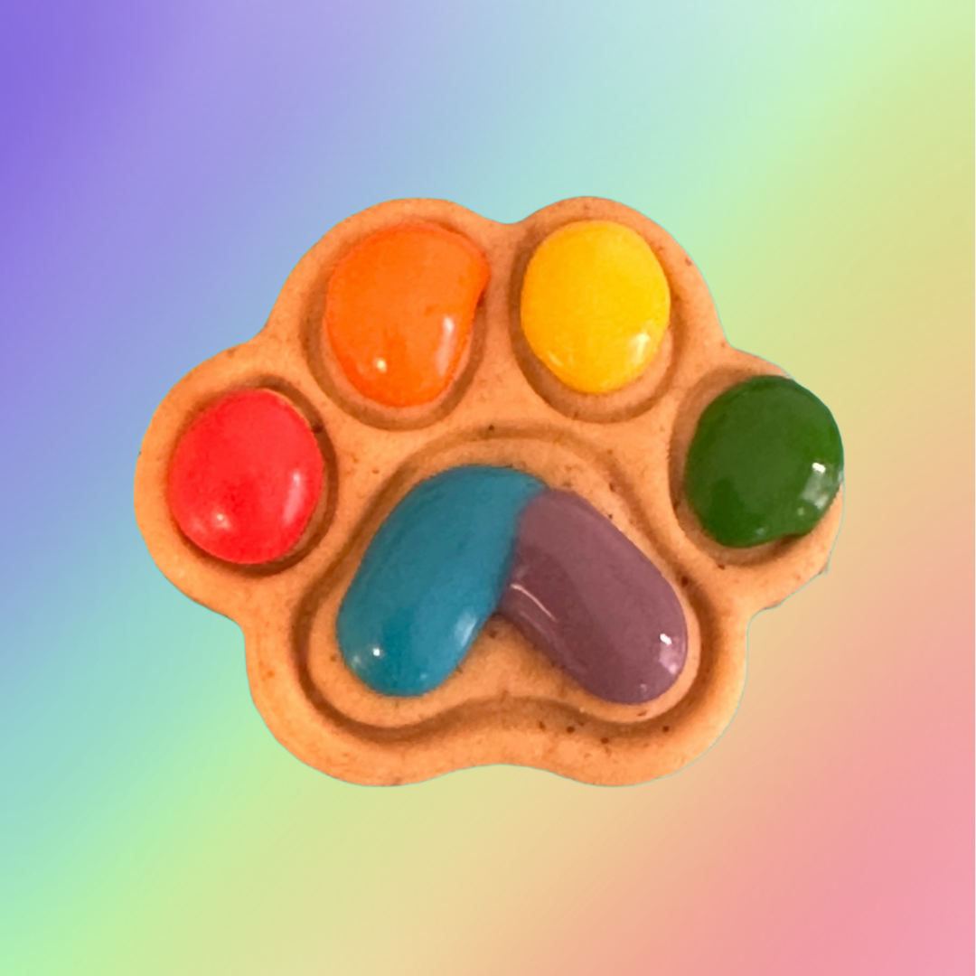A paw-shaped dog treat with multi-colored icing resembling a rainbow on a rainbow background.