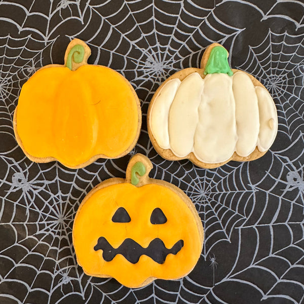 Three hand-decorated dog biscuits shaped like pumpkins, placed on a spider web background. One is a ghost pumpkin, one is a standard pumpkin, and one is a spooky pumpkin.