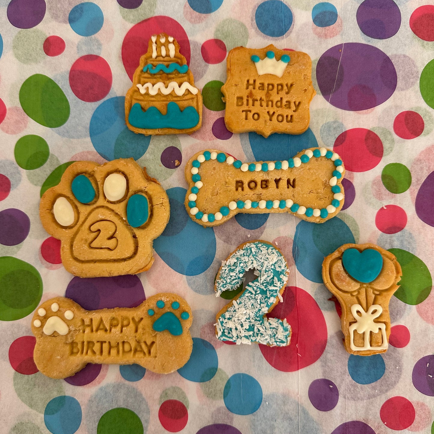 A selection of personalised dog biscuits in various shapes including a birthday cake, a paw print, a bone, and text messages saying 'Happy Birthday' and the age '2'. The biscuits are decorated with colourful icing and sprinkles.