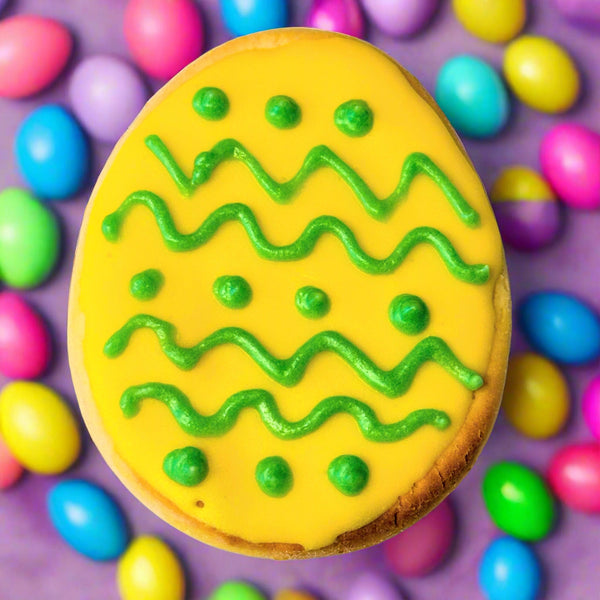 A dog treat shaped and decorated like an Easter egg, with yellow icing and green decorative lines, surrounded by colorful candy.
