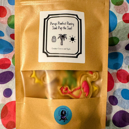 A packaged dog biscuit treat with a tropical theme, featuring graphics of palm trees and flamingos, and a label that reads 'Percy's Pawfect Pantry - Soak Pup The Sun'.
