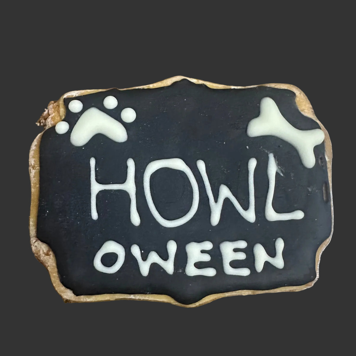 Large black iced Halloween dog biscuit with white “Howl-oween” lettering, paw print and bone decorations.