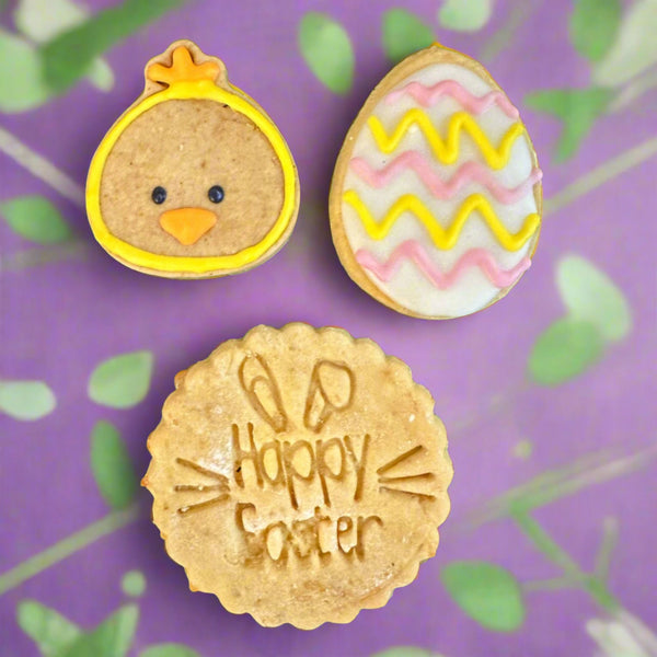 A set of three Easter-themed dog biscuits, with one shaped like an Easter egg, another like a chick, and a third with 'Happy Easter' text on it, all placed on a purple background with leaf patterns.
