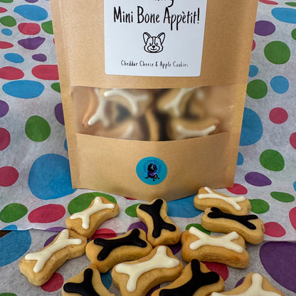 A treat bag labeled 'Mini Bone Appetit! Cheddar Cheese & Apple Cookies' with a transparent window showing mini bone-shaped dog treats inside. The treats are presented on a patterned background with a few treats scattered in front.