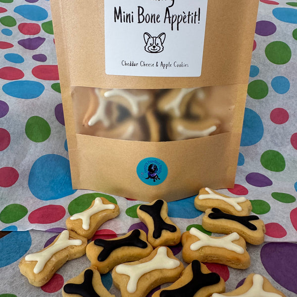 A treat bag labeled 'Mini Bone Appetit! Cheddar Cheese & Apple Cookies' with a transparent window showing mini bone-shaped dog treats inside. The treats are presented on a patterned background with a few treats scattered in front.
