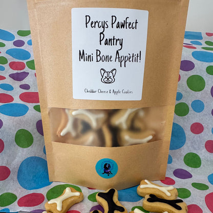 A treat bag labeled 'Mini Bone Appetit! Cheddar Cheese & Apple Cookies' with a transparent window showing mini bone-shaped dog treats inside. The treats are presented on a patterned background with a few treats scattered in front.