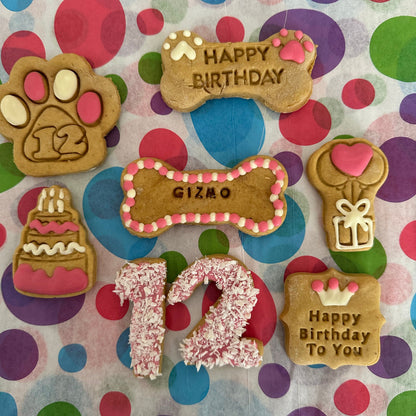A selection of personalised dog biscuits in various shapes including a birthday cake, a paw print, a bone, and text messages saying 'Happy Birthday' and the age '12'. The biscuits are decorated with colourful icing and sprinkles.