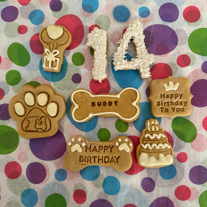 A selection of personalised dog biscuits in various shapes including a birthday cake, a paw print, a bone, and text messages saying 'Happy Birthday' and the age '14'. The biscuits are decorated with colourful icing and sprinkles.