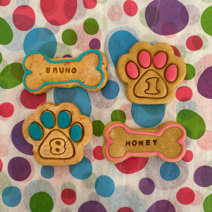 A selection of personalised dog biscuits in the shapes of bones and paws, decorated with icing in various colours, displayed on a polka dot background.