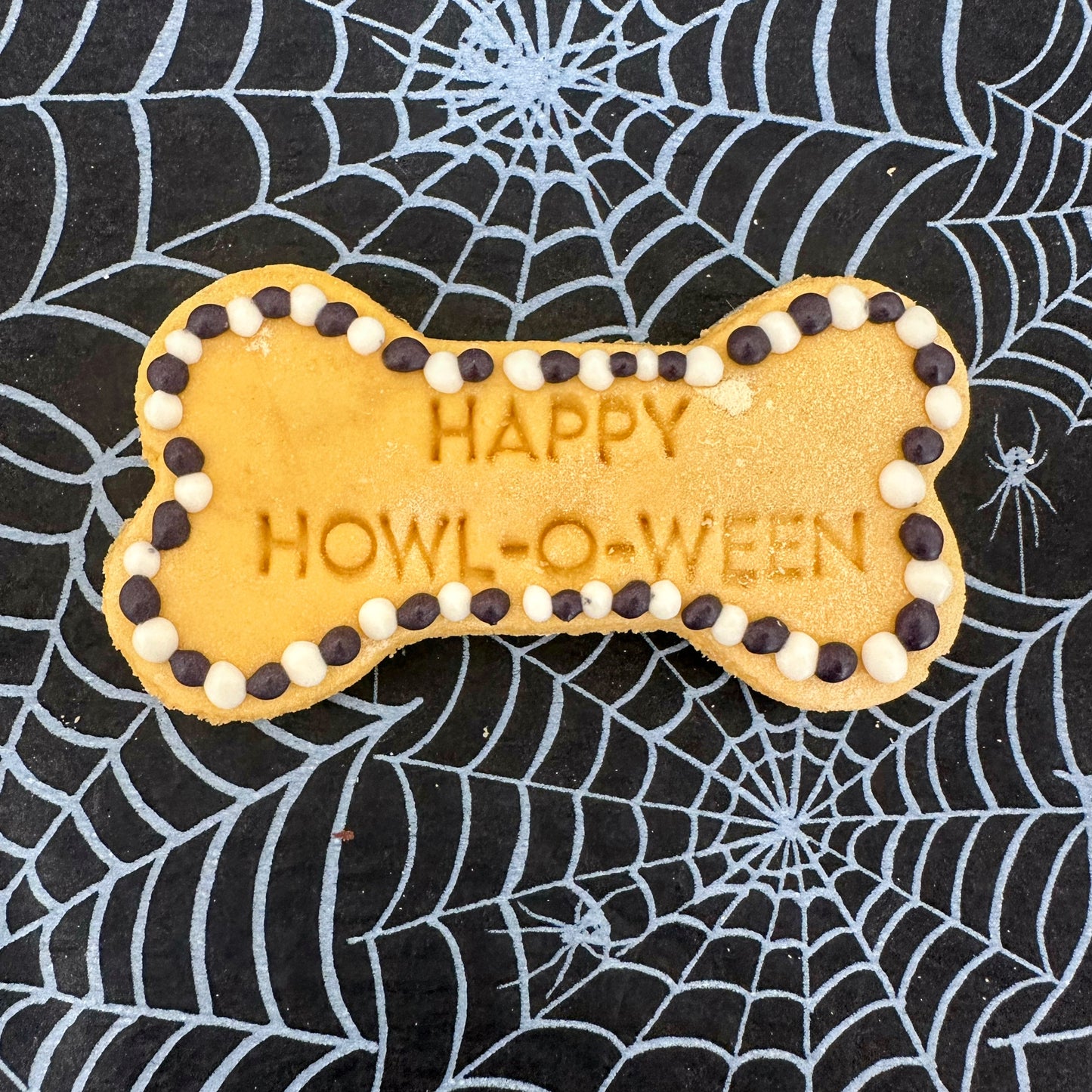 Dog bone-shaped cookie with 'Happy Howl-o-ween' text on a spider web background