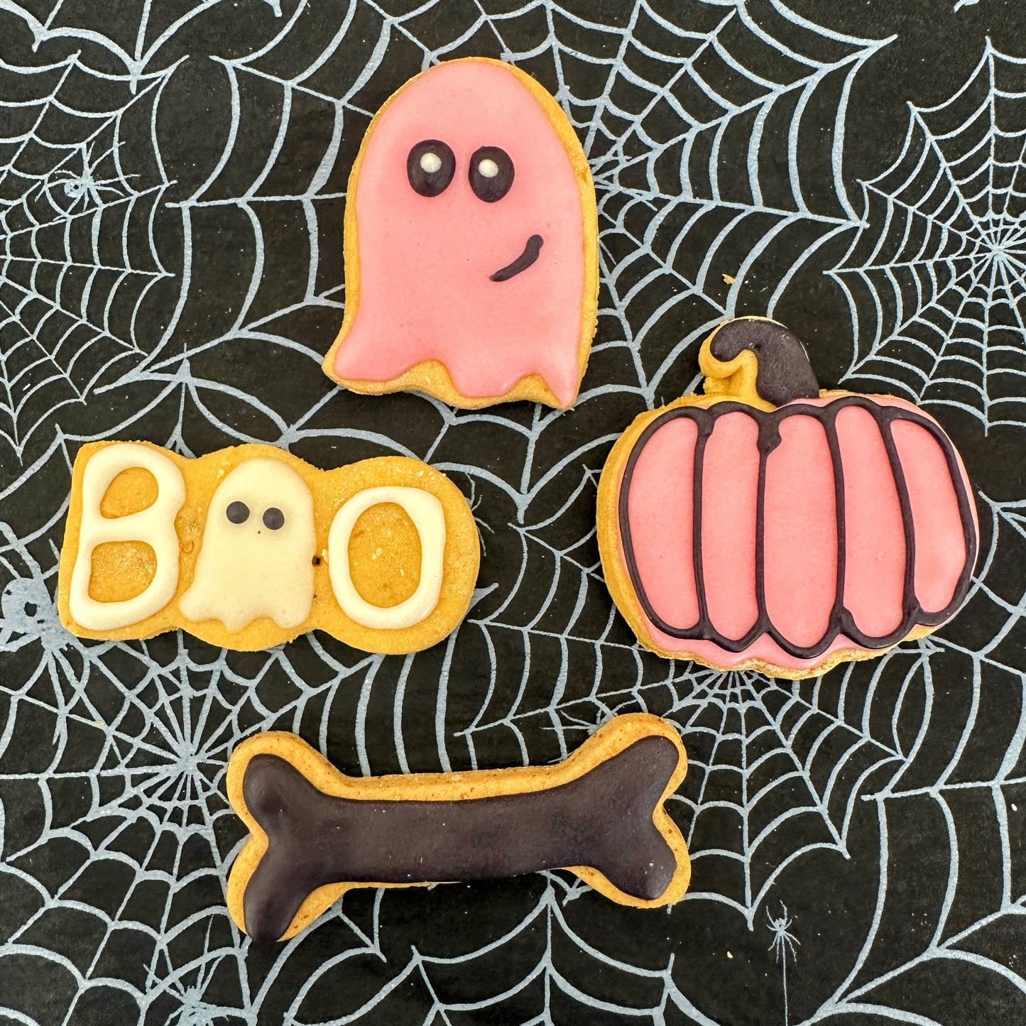 Halloween-themed cookies shaped like a ghost, pumpkin, 'BOO', and bone on a spider web background.