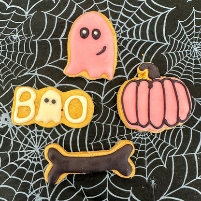 Halloween-themed cookies shaped like a ghost, pumpkin, 'BOO', and bone on a spider web background.