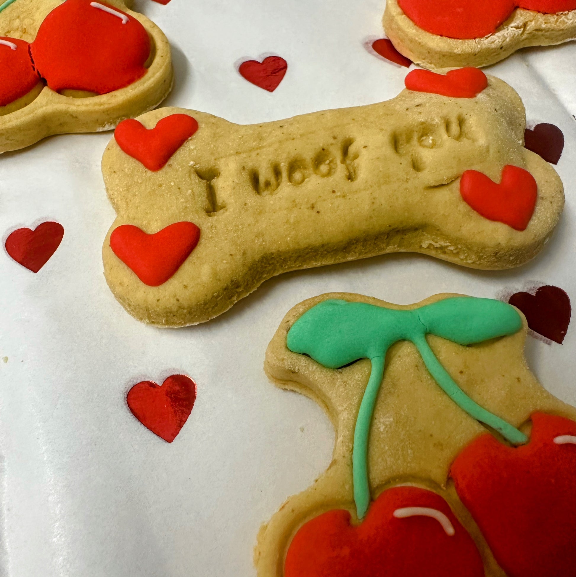 Valentine’s dog biscuits: a bone-shaped ‘I woof you’ biscuit with red hearts, surrounded by cute cherry-shaped biscuits with red icing, green tops.