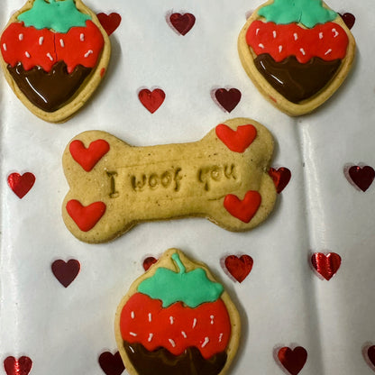 Valentine’s dog biscuits: a bone-shaped ‘I woof you’ biscuit with red hearts, surrounded by cute strawberry-shaped biscuits with red icing, green tops, and chocolate-dipped bottoms