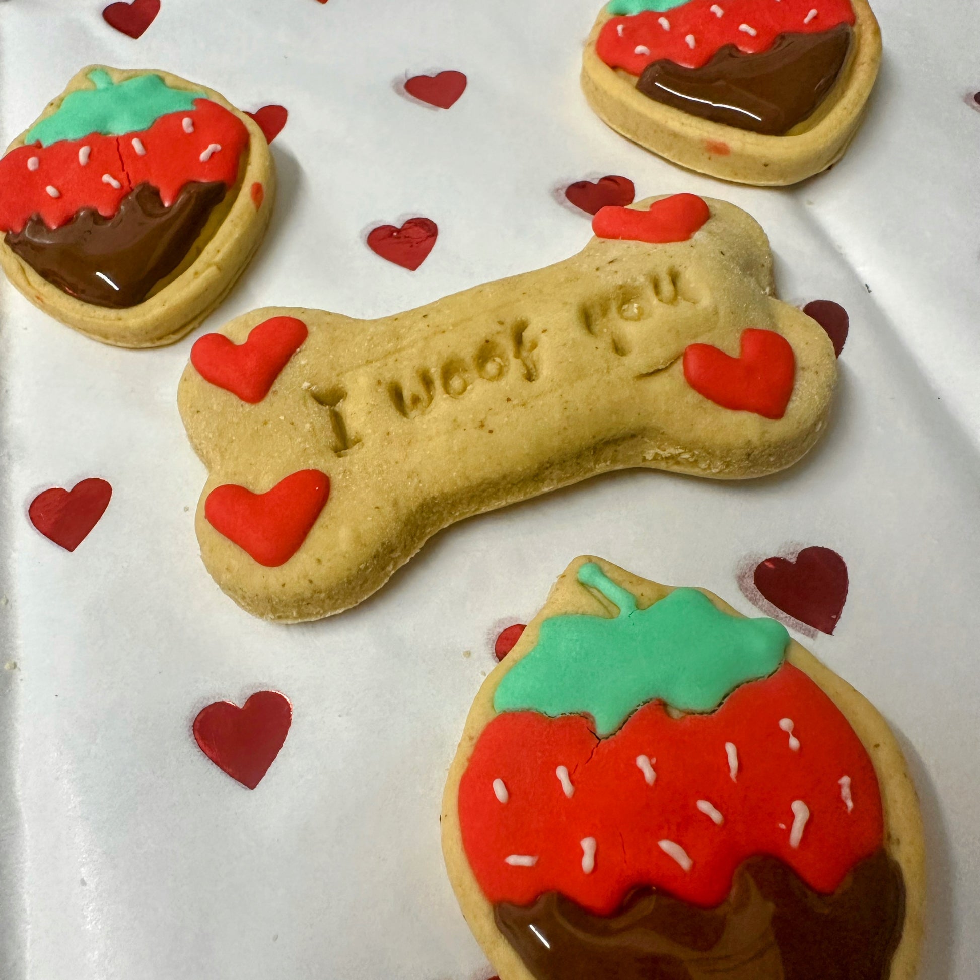 A set of Valentine’s-themed dog biscuits on white paper with small red heart decorations. In the center is a bone-shaped biscuit stamped with 'I woof you' and decorated with four small red icing hearts. Surrounding it are strawberry-shaped biscuits with red icing, green tops, and a chocolate-dipped bottom