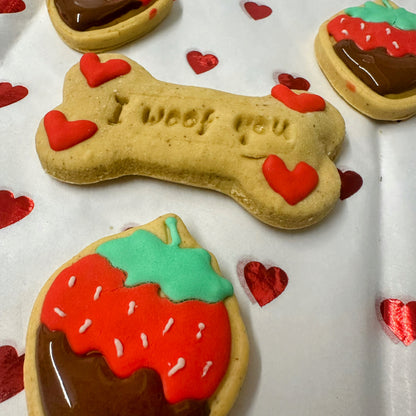 Valentine’s dog biscuits: a bone-shaped ‘I woof you’ biscuit with red hearts, surrounded by cute strawberry-shaped biscuits with red icing, green tops, and chocolate-dipped bottoms