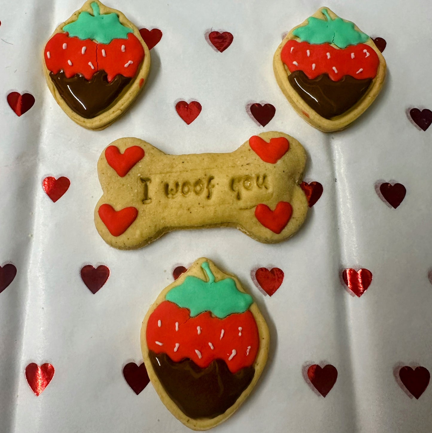 Dog bone-shaped cookie with 'I woof you' text, surrounded by strawberry-shaped cookies and red hearts on a white background.