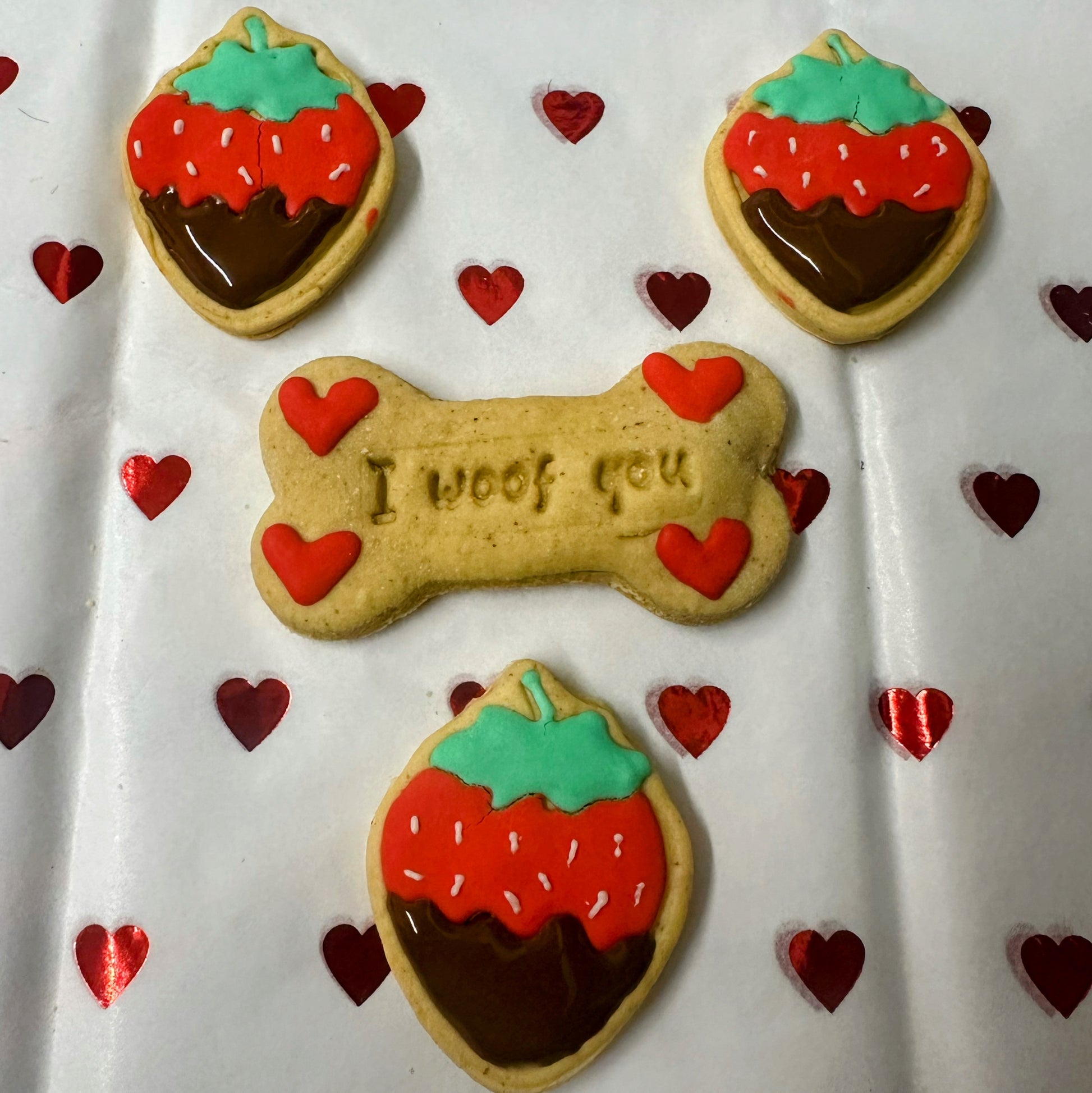 Valentine’s dog biscuits: a bone-shaped ‘I woof you’ biscuit with red hearts, surrounded by cute strawberry-shaped biscuits with red icing, green tops, and chocolate-dipped bottoms