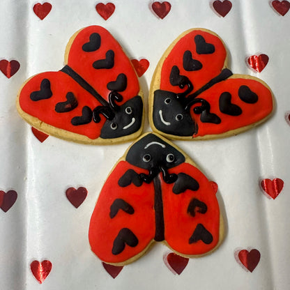 Three ladybug-shaped cookies with red bodies, black spots, and black heads on a white background with red heart patterns.