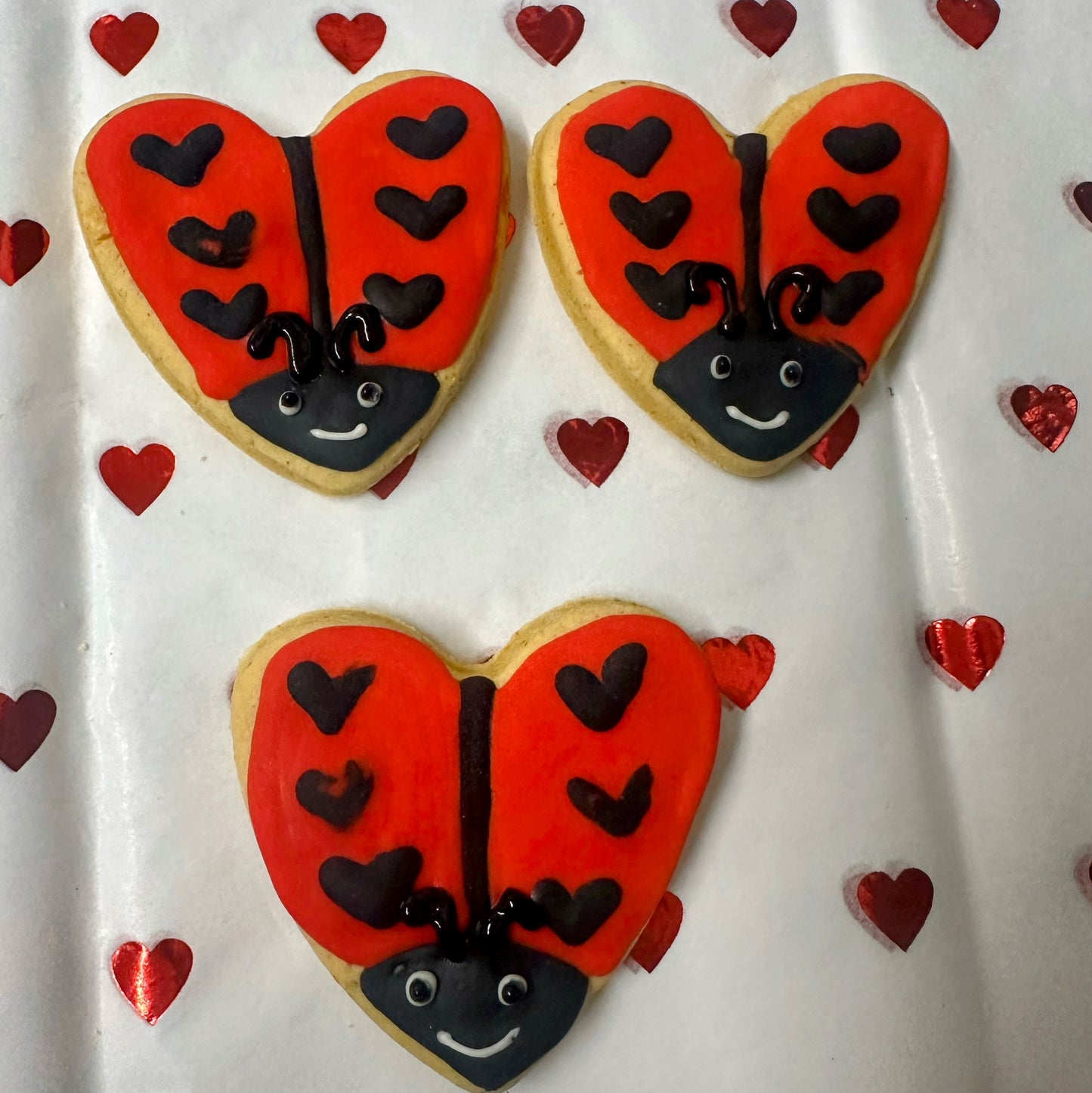 Heart-shaped cookie decorated like a ladybug with red wings and black spots on a white background with small red hearts.
