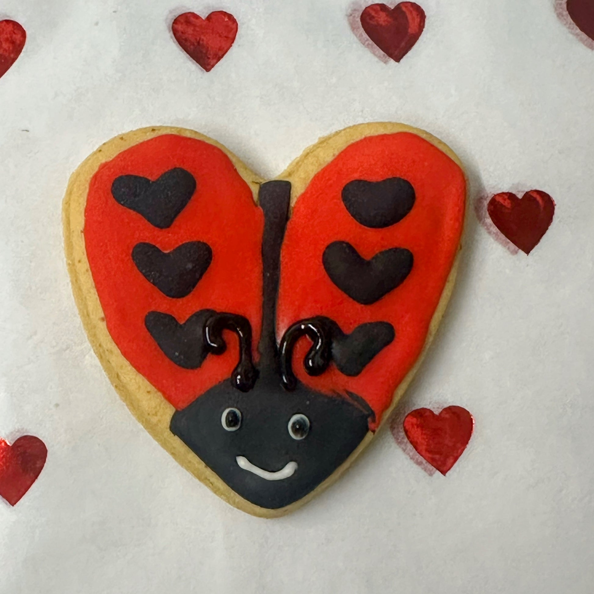 Heart-shaped cookie decorated like a ladybug with red wings and black spots on a white background with small red hearts.