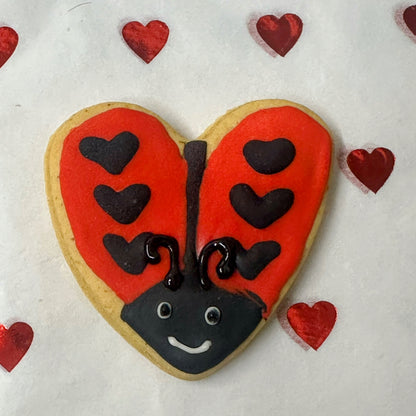 Heart-shaped cookie decorated like a ladybug with red wings, black body, and face on a white background with small red hearts.