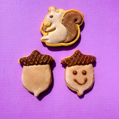 Three decorated cookies shaped like a squirrel and two acorns on a purple background