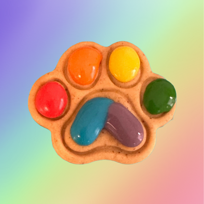 A paw-shaped dog treat with multi-colored icing resembling a rainbow on a rainbow background.
